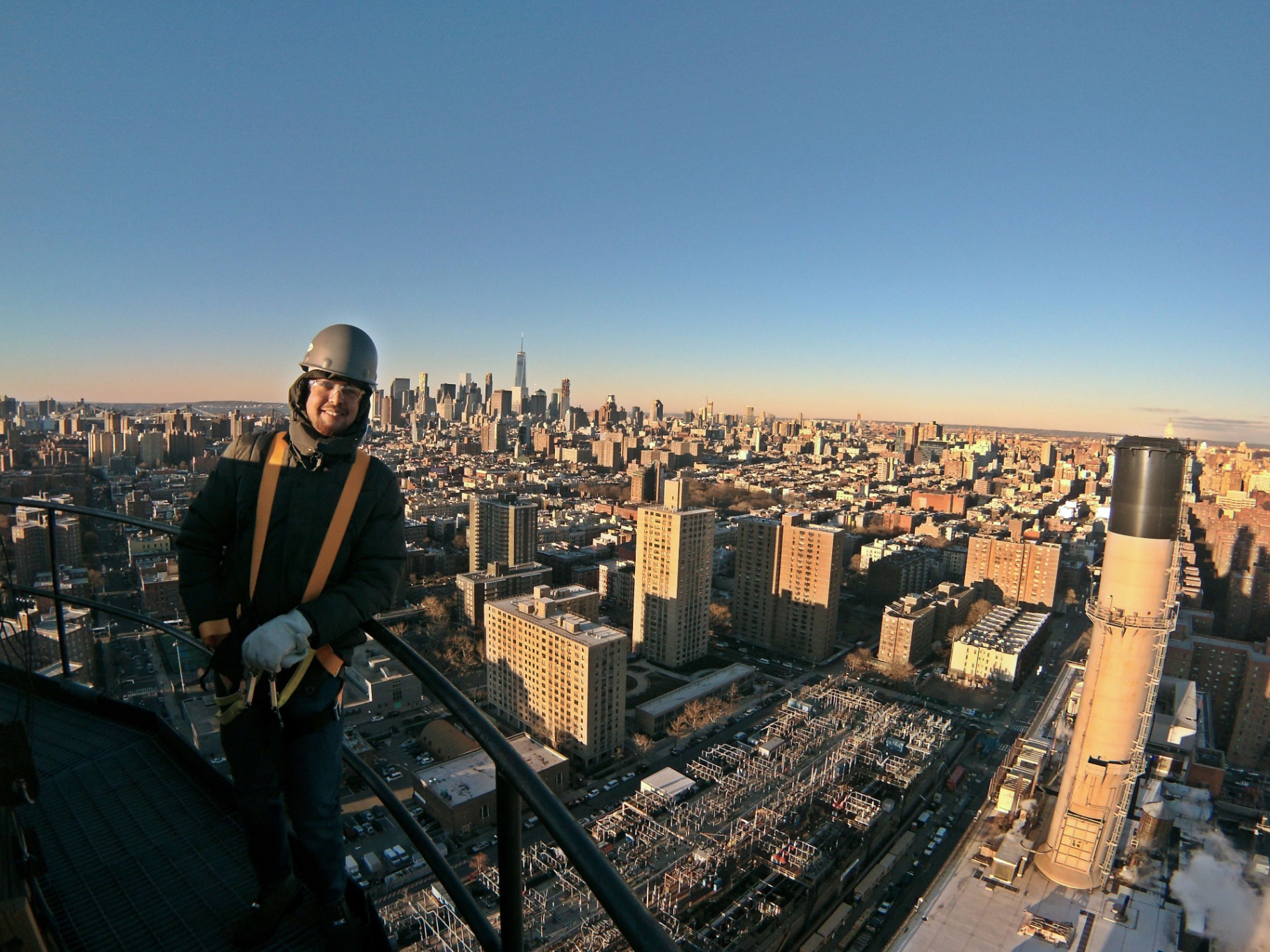Ali Akbar in hardhat, safety glasses, and rigging harness on top of Con Edison's East River Generating Station in Manhattan, with the Lower Manhattan skyline behind him.